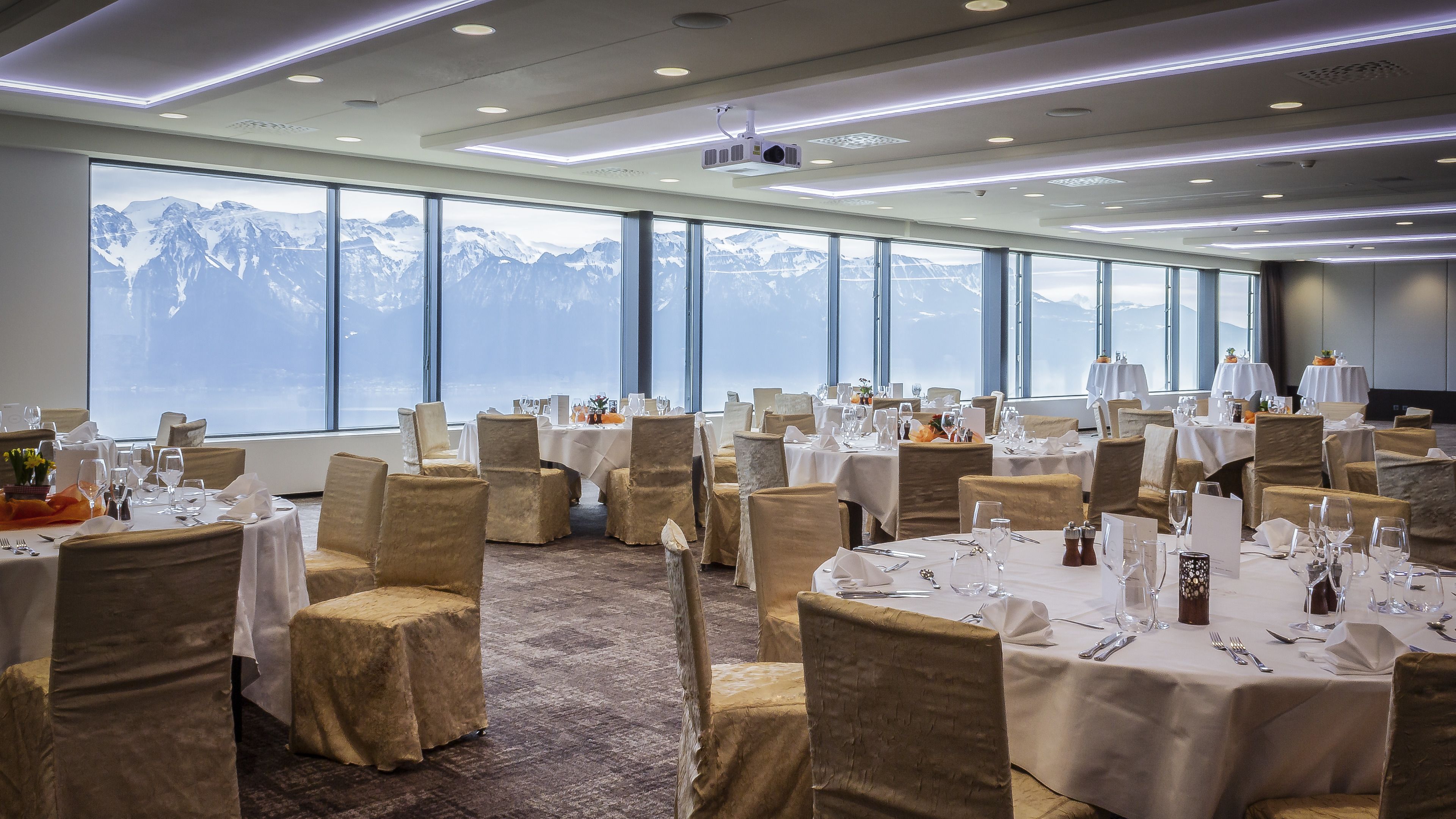 Ein Festsaal mit gedeckten Tischen, großen Fenstern mit Bergblick und einem modernen Deckenstil.