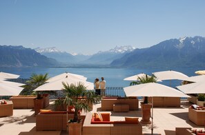 Een terras met rieten meubilair en parasols, met uitzicht op een meer en bergen op de achtergrond.
