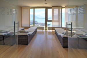 Quarto de hotel moderno com duas áreas de estar distintas, amplas janelas com vista para a cidade e para o lago, e piso de madeira.