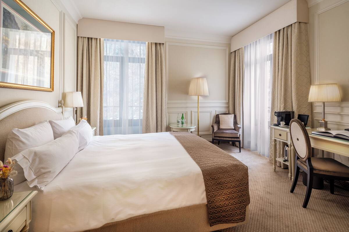 Ein Hotelzimmer mit einem großen Bett, einem Schreibtisch, einem Stuhl, einer Lampe und einem Vorhangfenster.