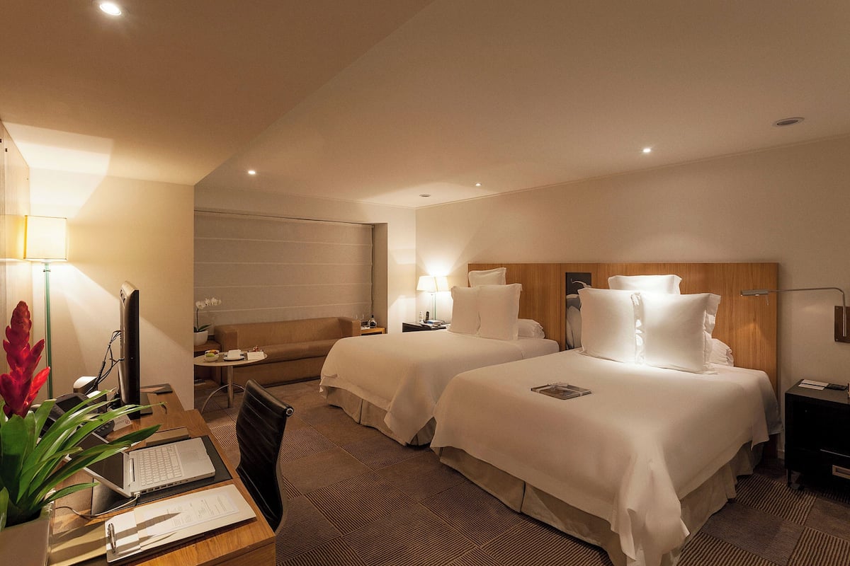 Ein Hotelzimmer mit zwei Betten, einem Schreibtisch mit Laptop, einem Stuhl, einem Nachttisch mit Lampe und einem großen Fenster.
