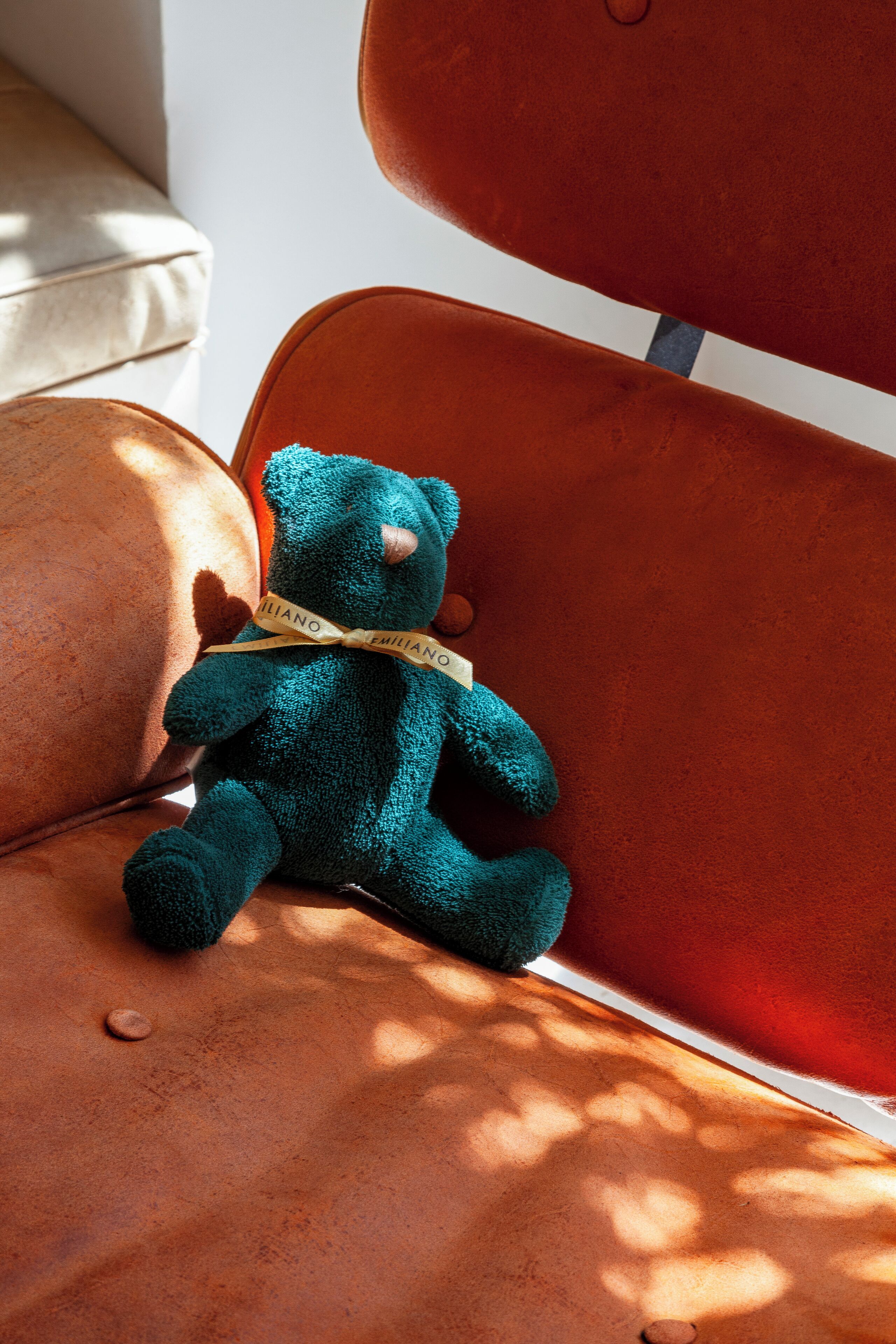 Um urso de pelúcia azul-esverdeado sentado em um sofá de couro marrom com uma fita no pescoço.