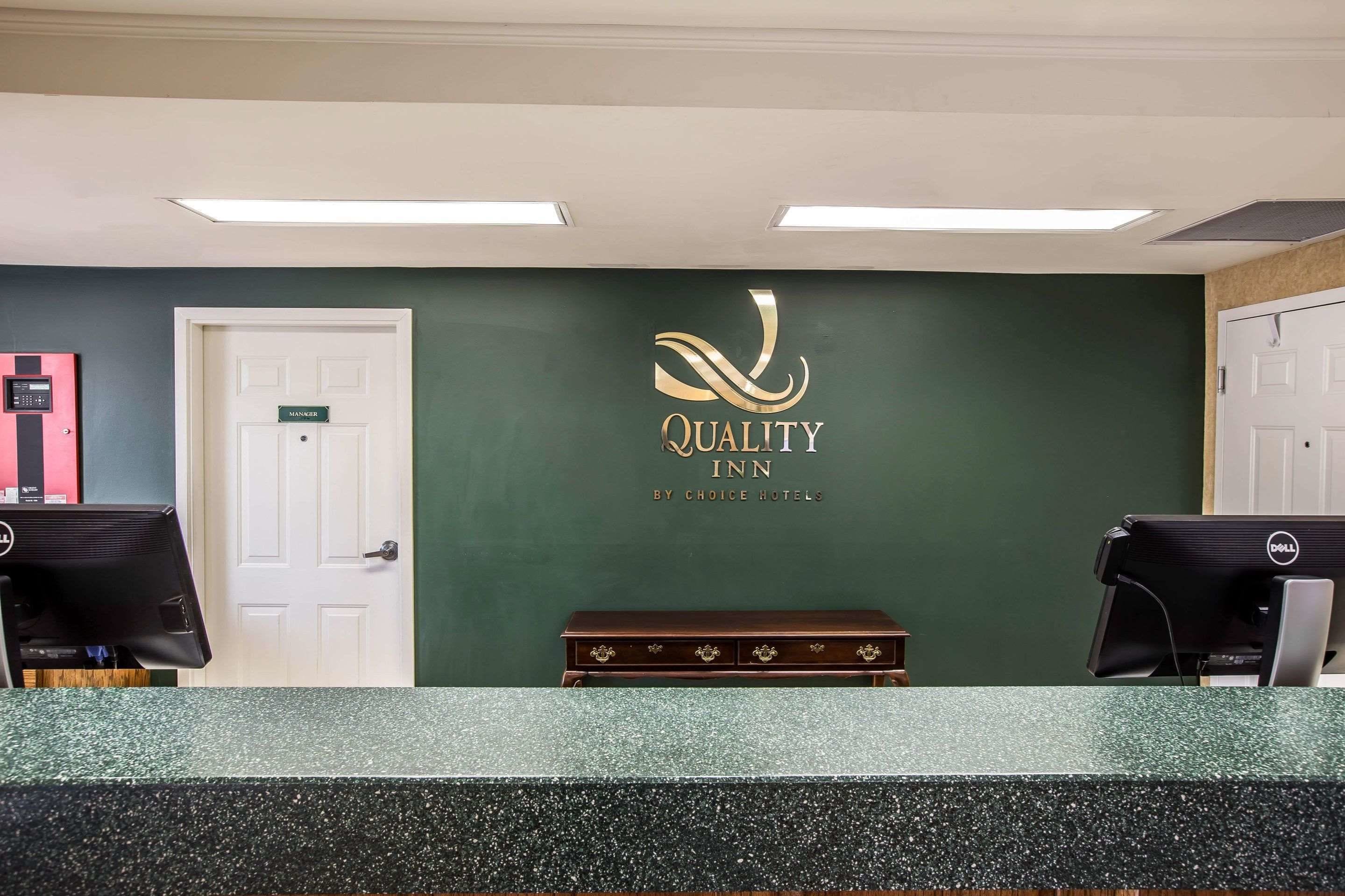 A reception desk with a green wall, a sign reading "Quality Inn by Choice Hotels," and two computer monitors.