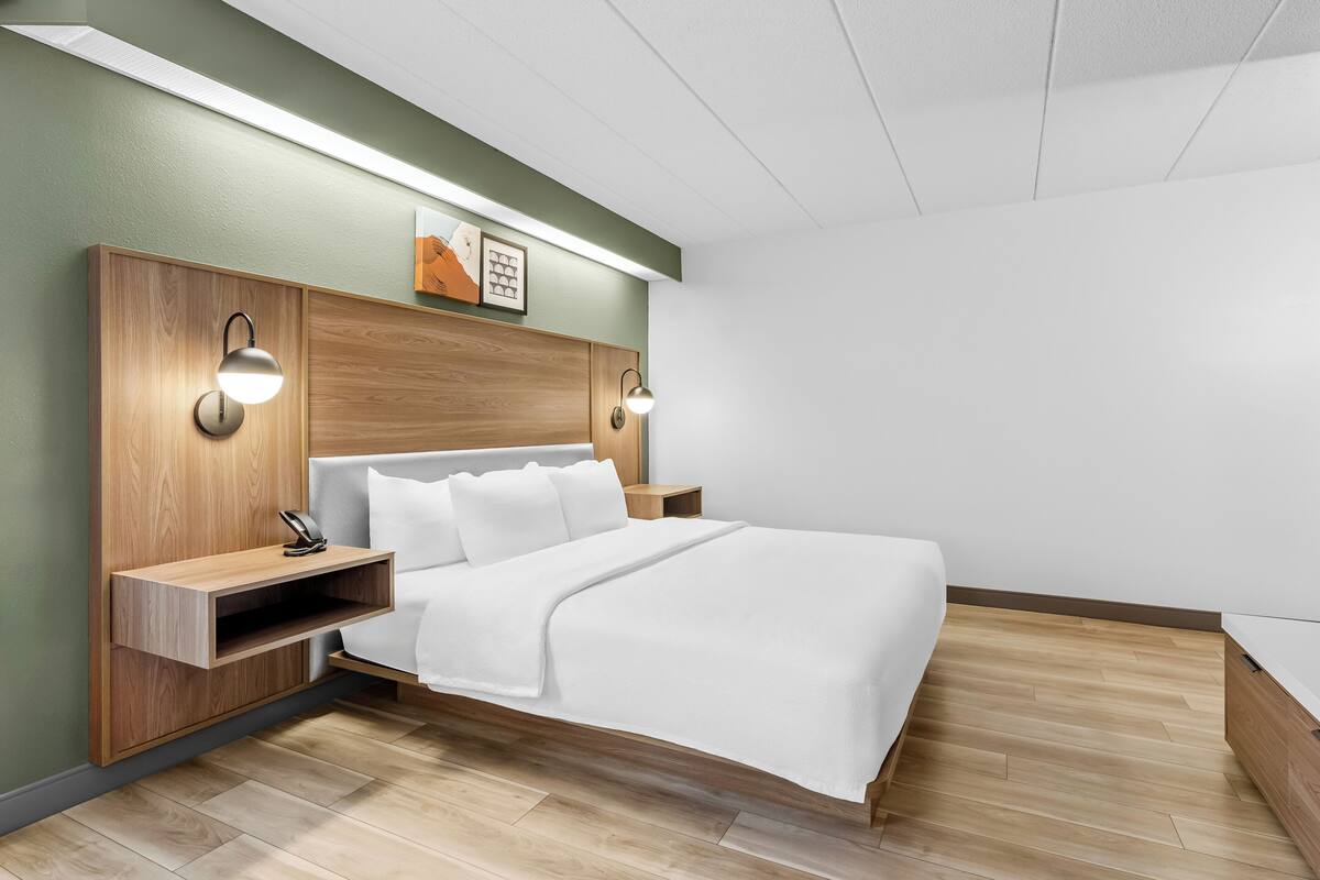 Une chambre d’hôtel moderne avec une tête de lit en bois, un lit avec une literie blanche, deux tables de chevet avec des lampes et une petite étagère.