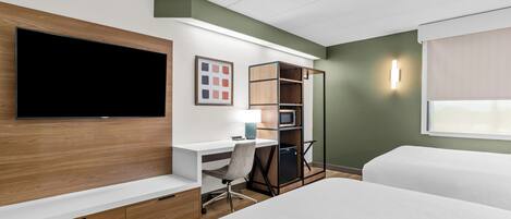 A hotel room with a flat-screen TV, a desk with a chair, a bookshelf, a bed, and a window with a curtain.