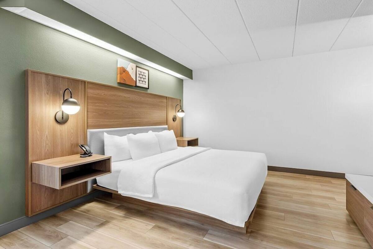 Une chambre d’hôtel moderne avec une tête de lit en bois, un lit blanc, deux tables de chevet avec des lampes et une étagère.