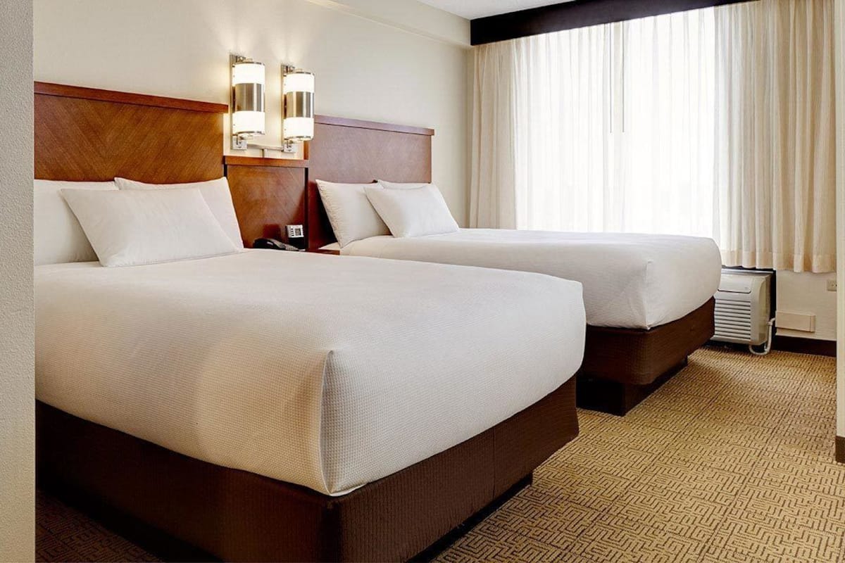 A hotel room with two beds, a wooden headboard, wall-mounted lights, a window with curtains, and a carpeted floor.