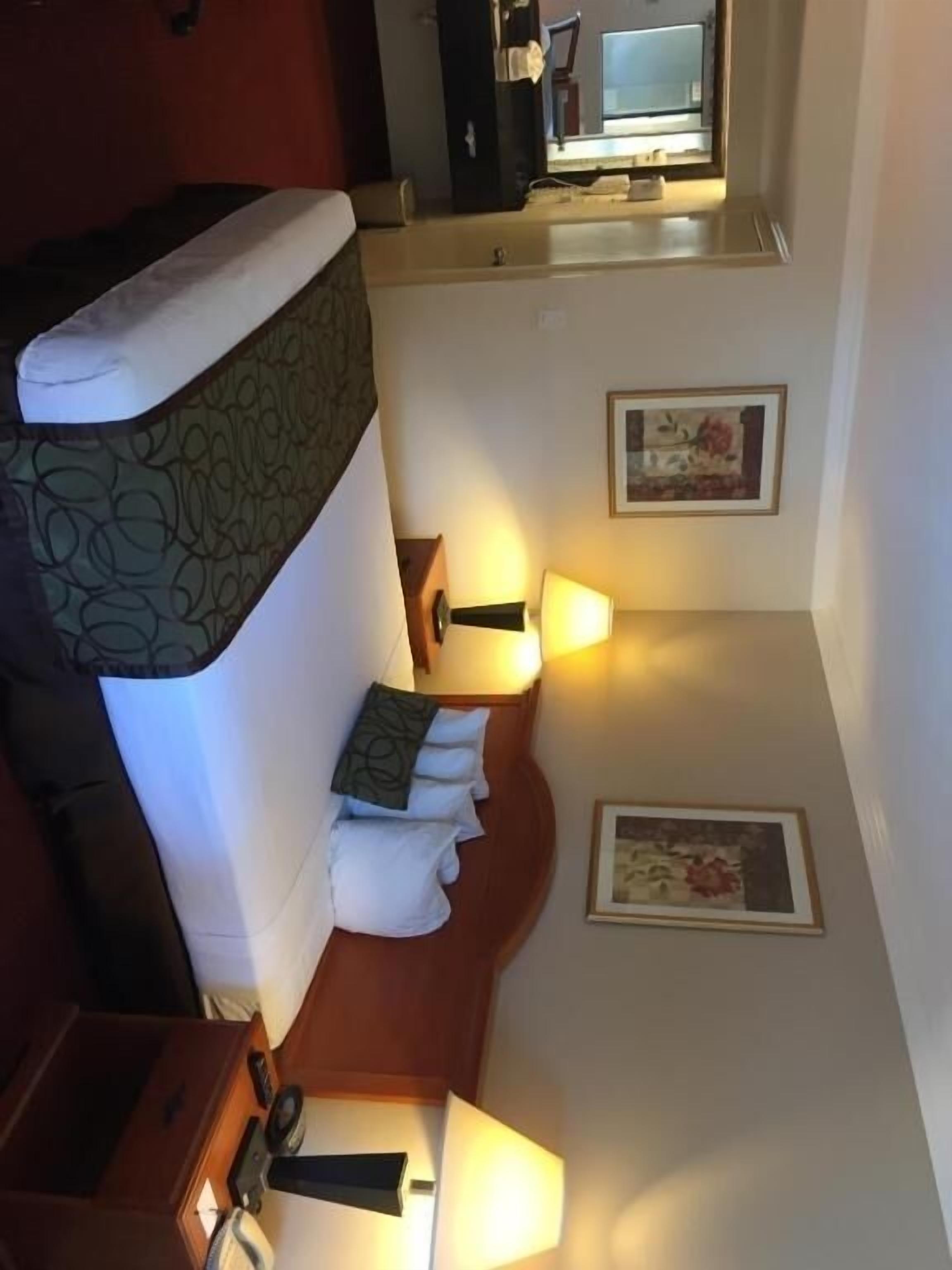 A hotel room with a bed, bedside lamps, a framed picture on the wall, and a door leading to another room.