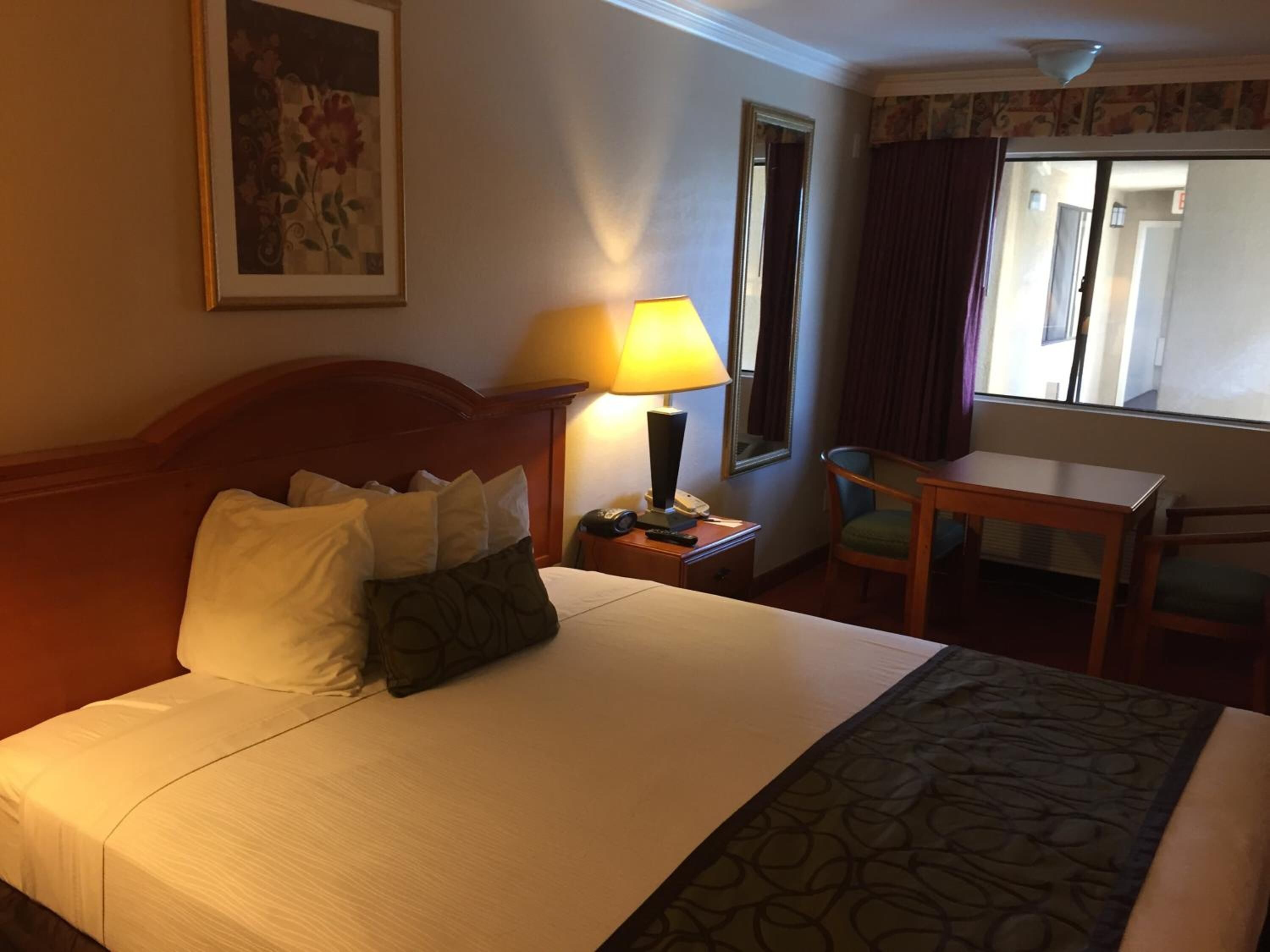 A hotel room with a bed, a nightstand with a lamp, a chair, a table, and a window with curtains.