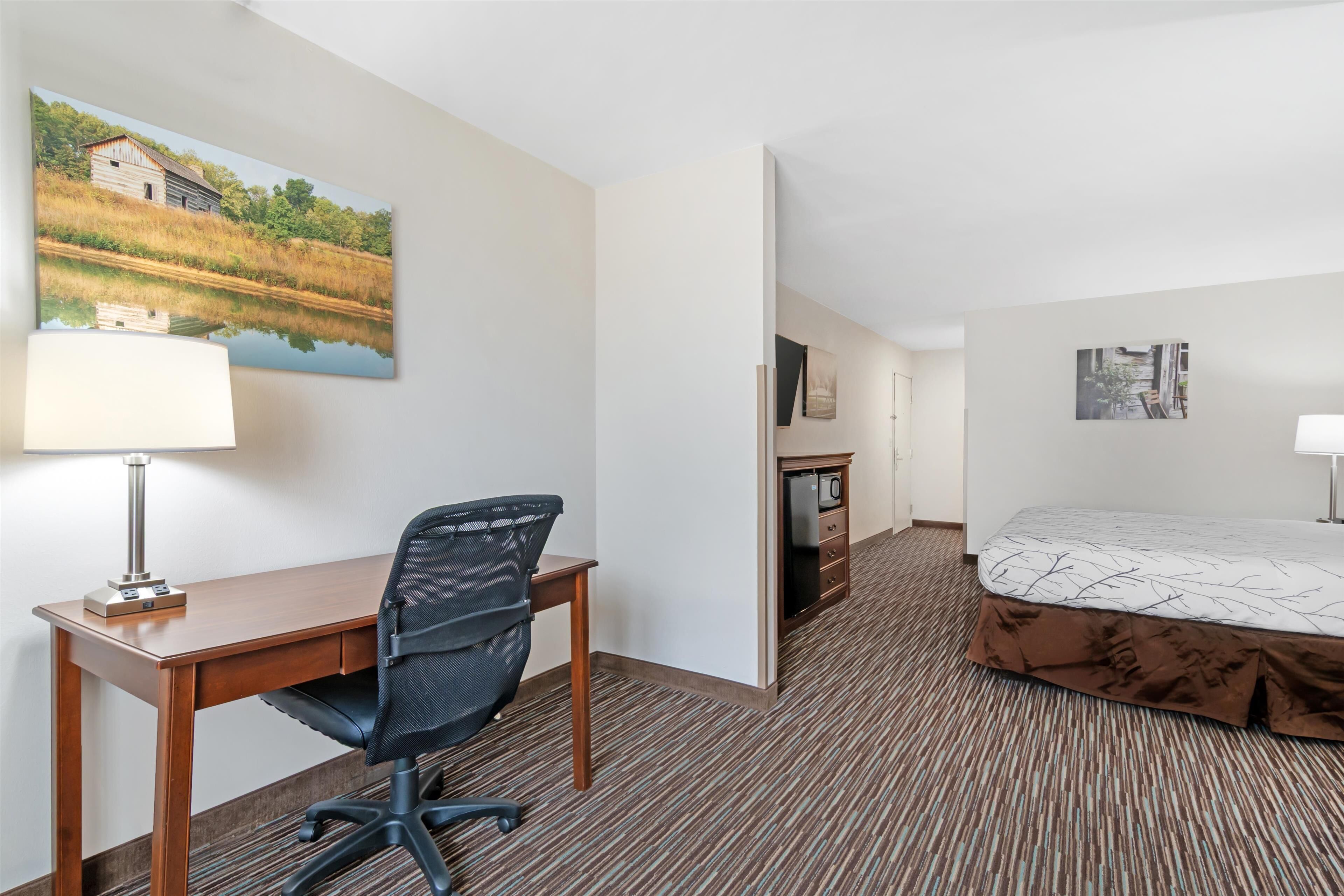 Ein Hotelzimmer mit einem Holztisch, einem schwarzen Bürostuhl, einem Bett mit gemustertem Überwurf, einem Wandbild von einem Haus am See und einem Teppichboden.