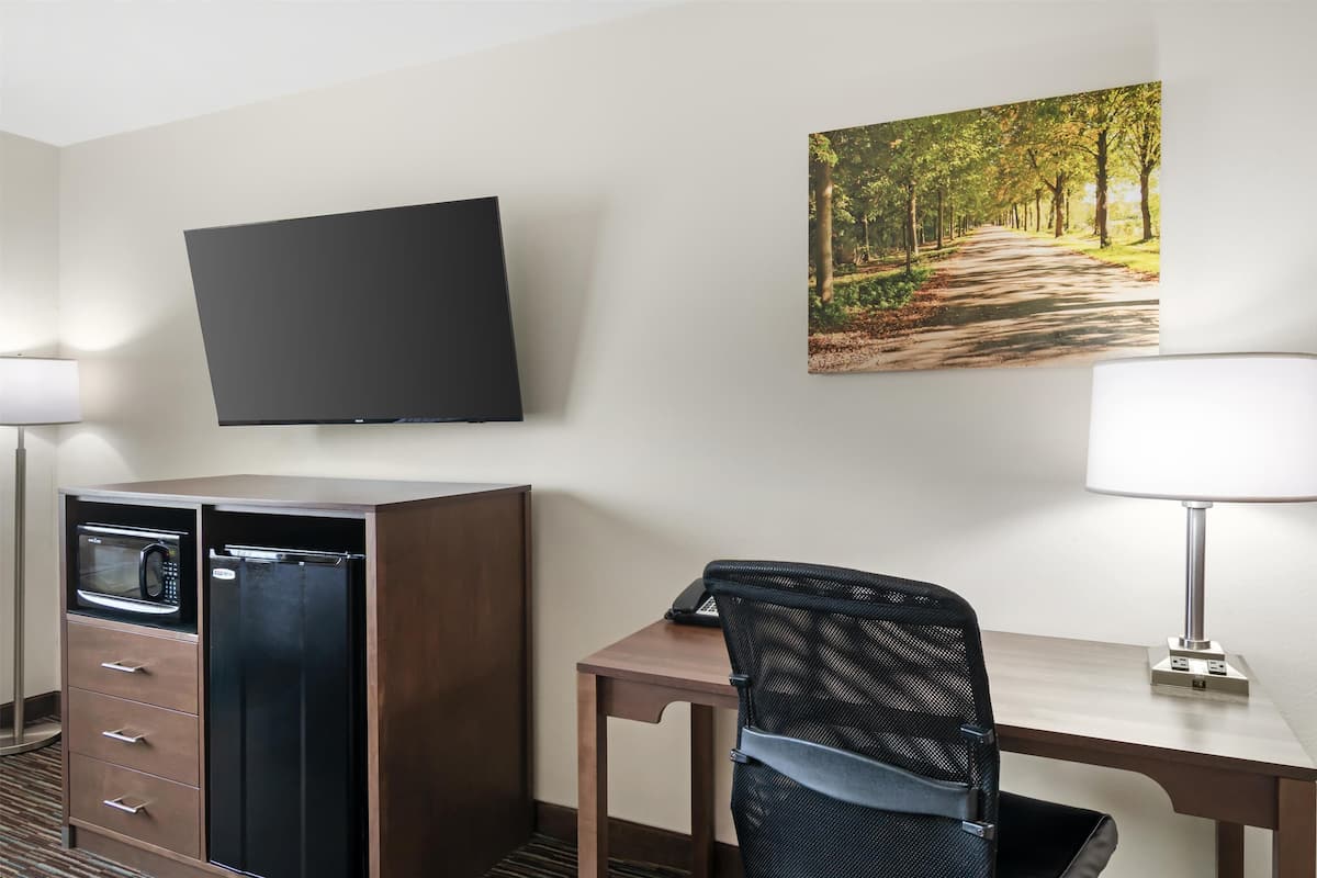 A hotel room with a flat-screen TV mounted on the wall, a desk with a chair, a microwave, and a refrigerator.