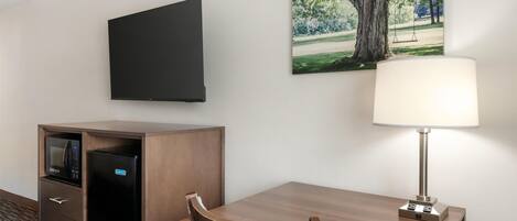 Habitación de hotel con televisor de pantalla plana montado en la pared, escritorio con lámpara, microondas y refrigerador en un mueble de madera.