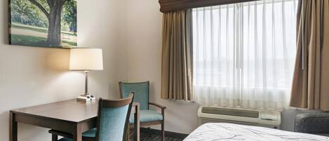 Une chambre d’hôtel avec un lit, un bureau et une chaise, une lampe, une fenêtre avec des rideaux et un tableau au mur.