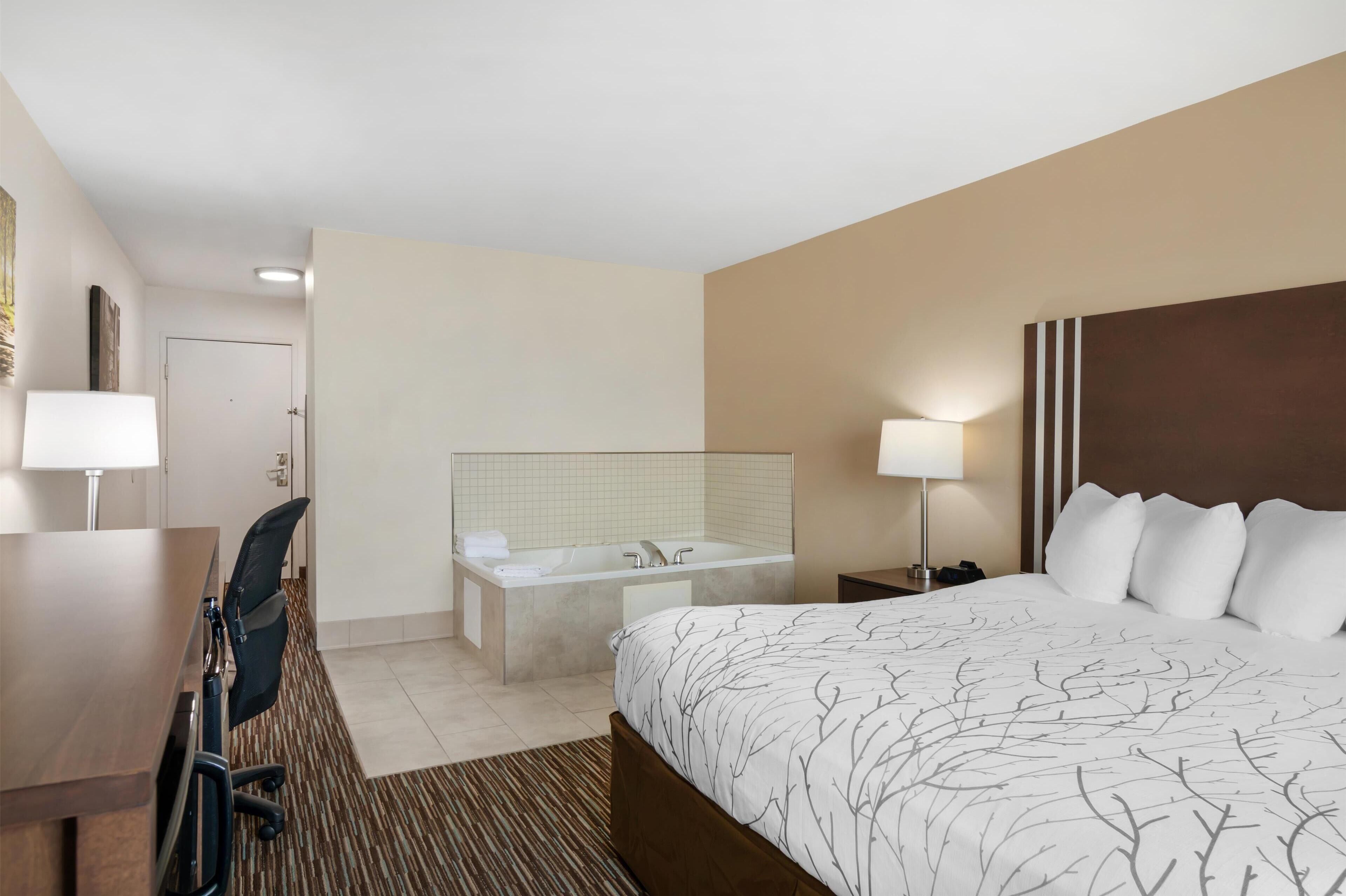 A hotel room with a bed, a desk, a chair, a bathtub, and a nightstand with a lamp.