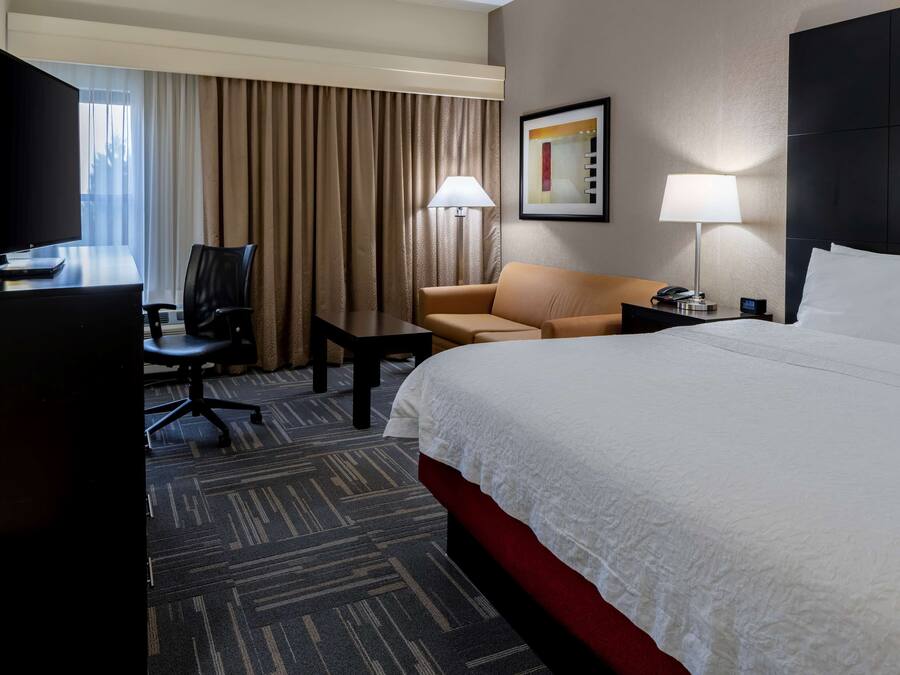 Habitación de hotel con una cama grande, un escritorio con televisor, una silla, una lámpara y un cuadro en la pared.