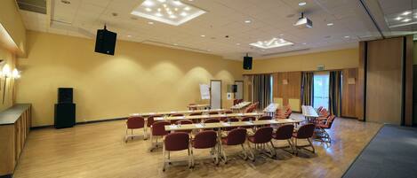 A conference room with wooden floors, rows of tables and chairs, a projector, and a whiteboard.