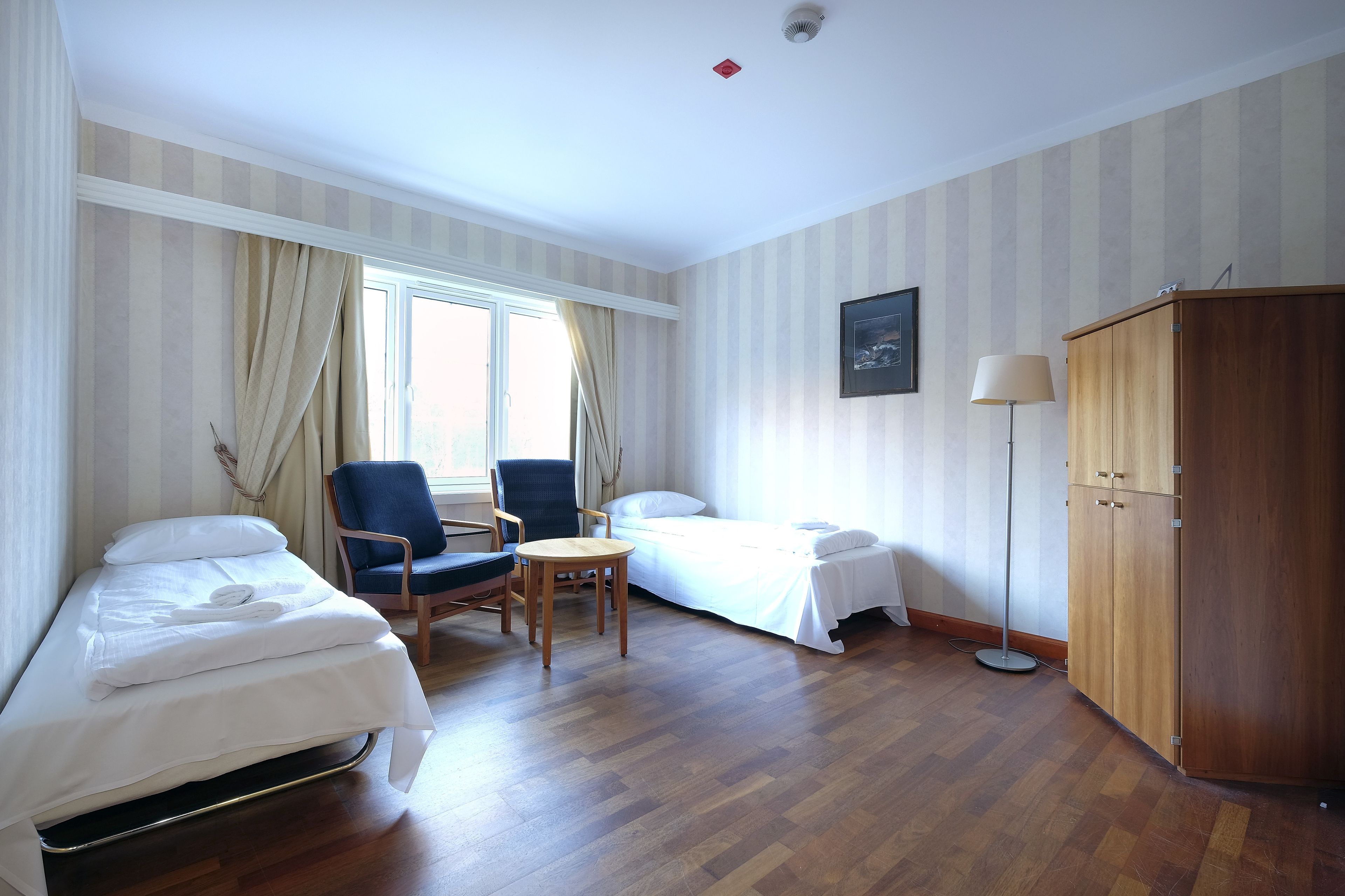 A hotel room with two single beds, a small table, a chair, a wardrobe, and a window with curtains.