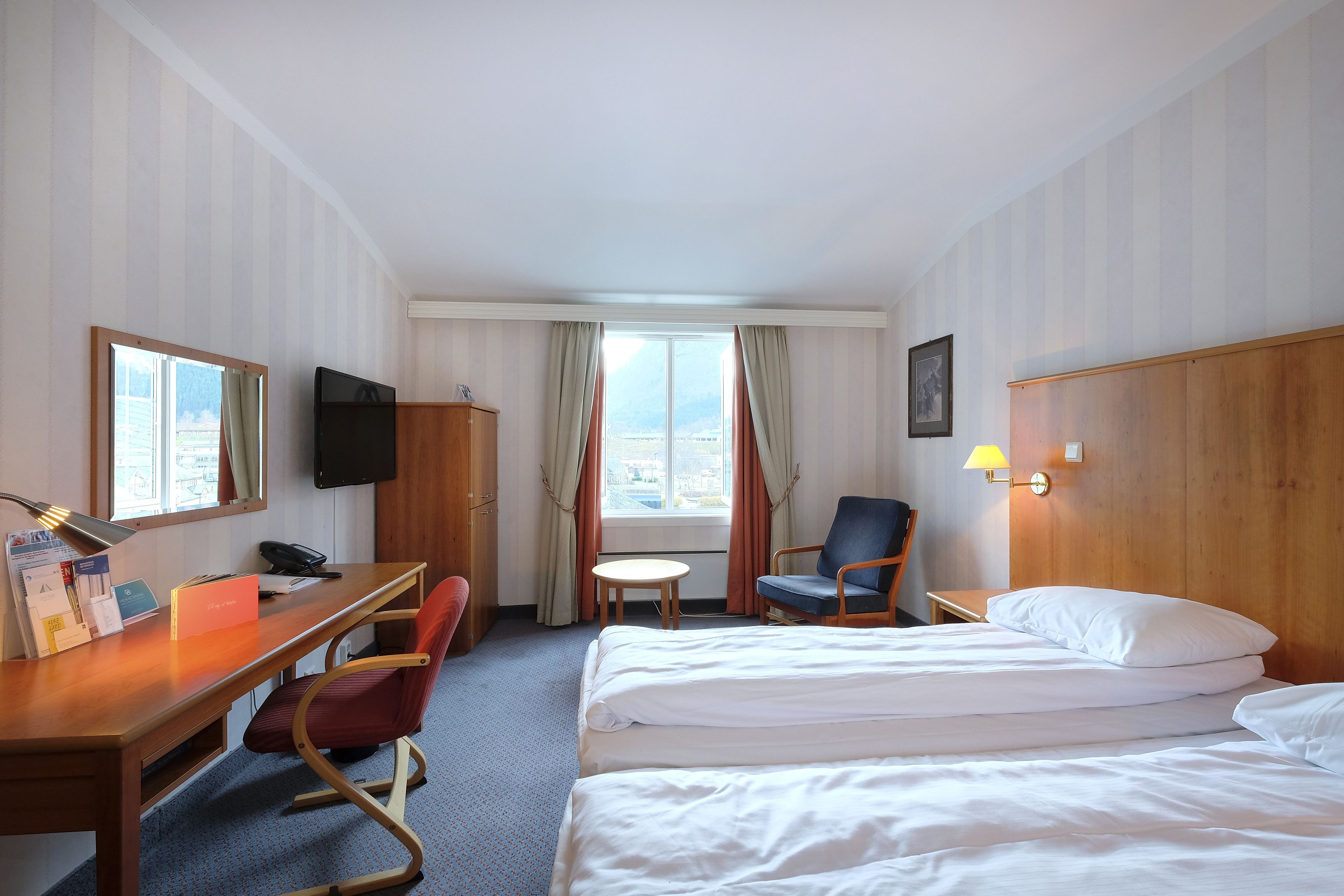 Habitación de hotel con cama, escritorio, silla, televisor y ventana con cortinas.
