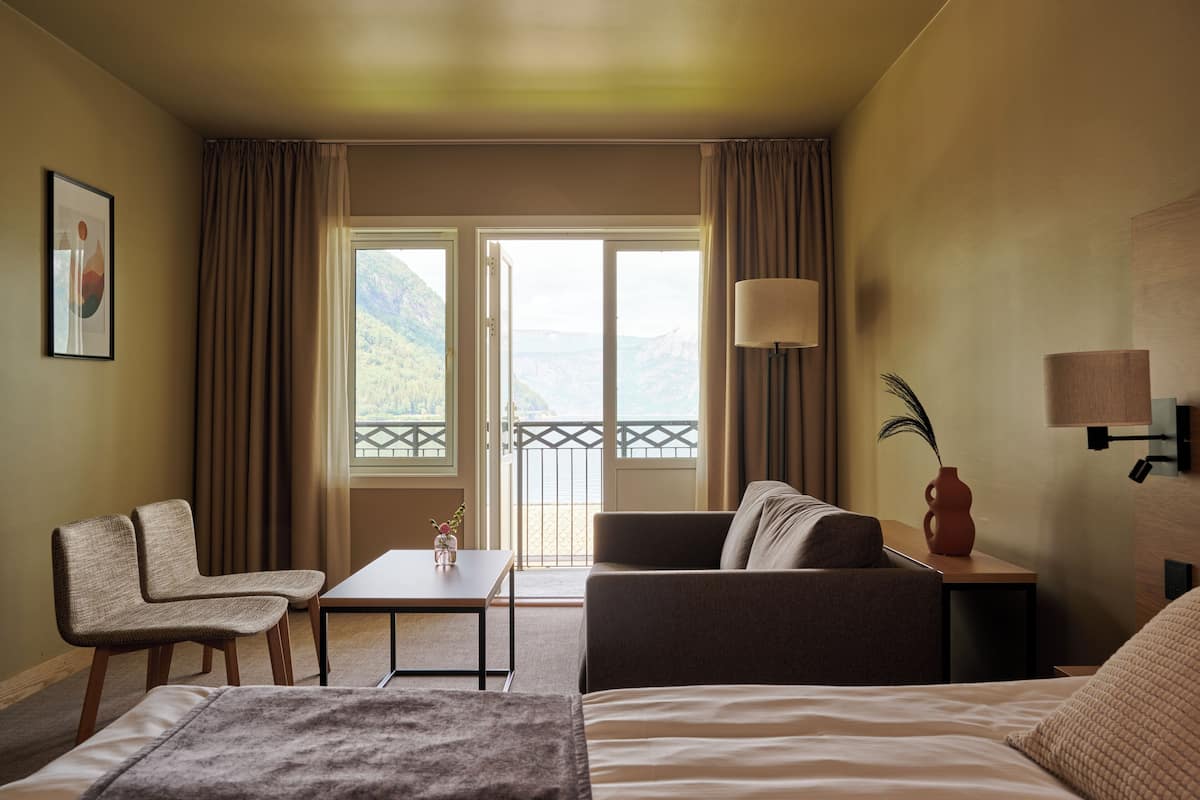 Une chambre d’hôtel avec un lit, deux fauteuils, un canapé, une table et une lampe. Il y a une fenêtre avec des rideaux et une vue sur les montagnes.