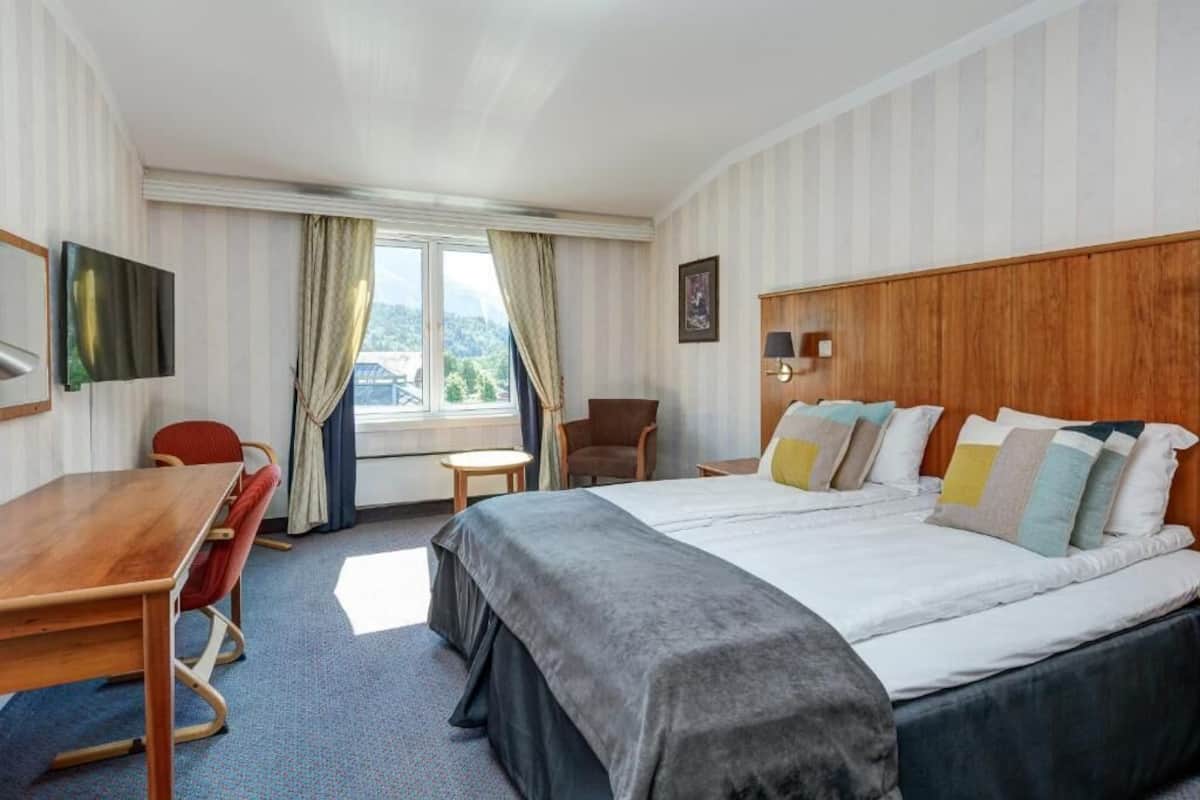 Une chambre d’hôtel comprenant un lit, un bureau, une chaise, une télévision, une fenêtre avec des rideaux et une vue sur les montagnes.