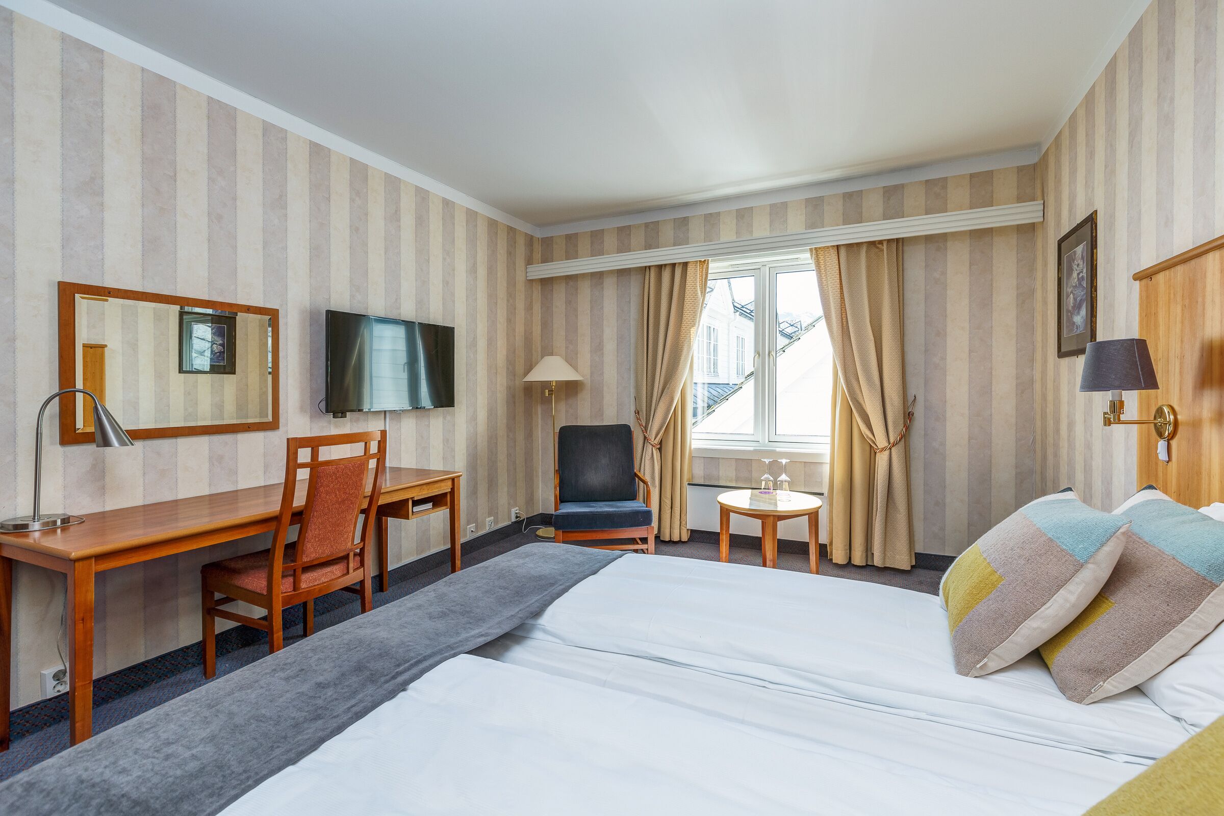 A hotel room with a bed, a desk, a chair, a TV, a lamp, and a window with curtains.