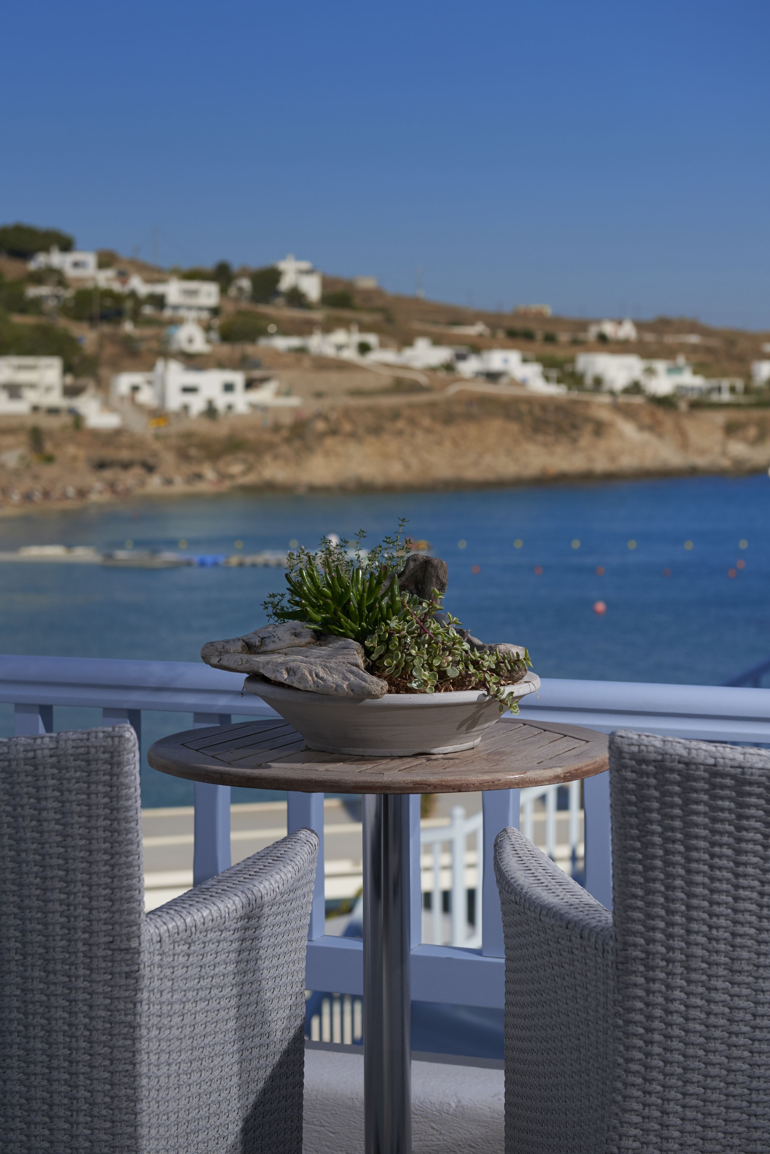 A round table with a potted plant on a balcony overlooking a coastal area with buildings in the background.
