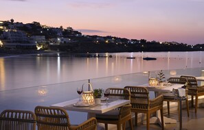 An outdoor dining area with wicker chairs and tables, a view of the water, and a cityscape in the background. - Petinos Beach Hotel (Mykonos)