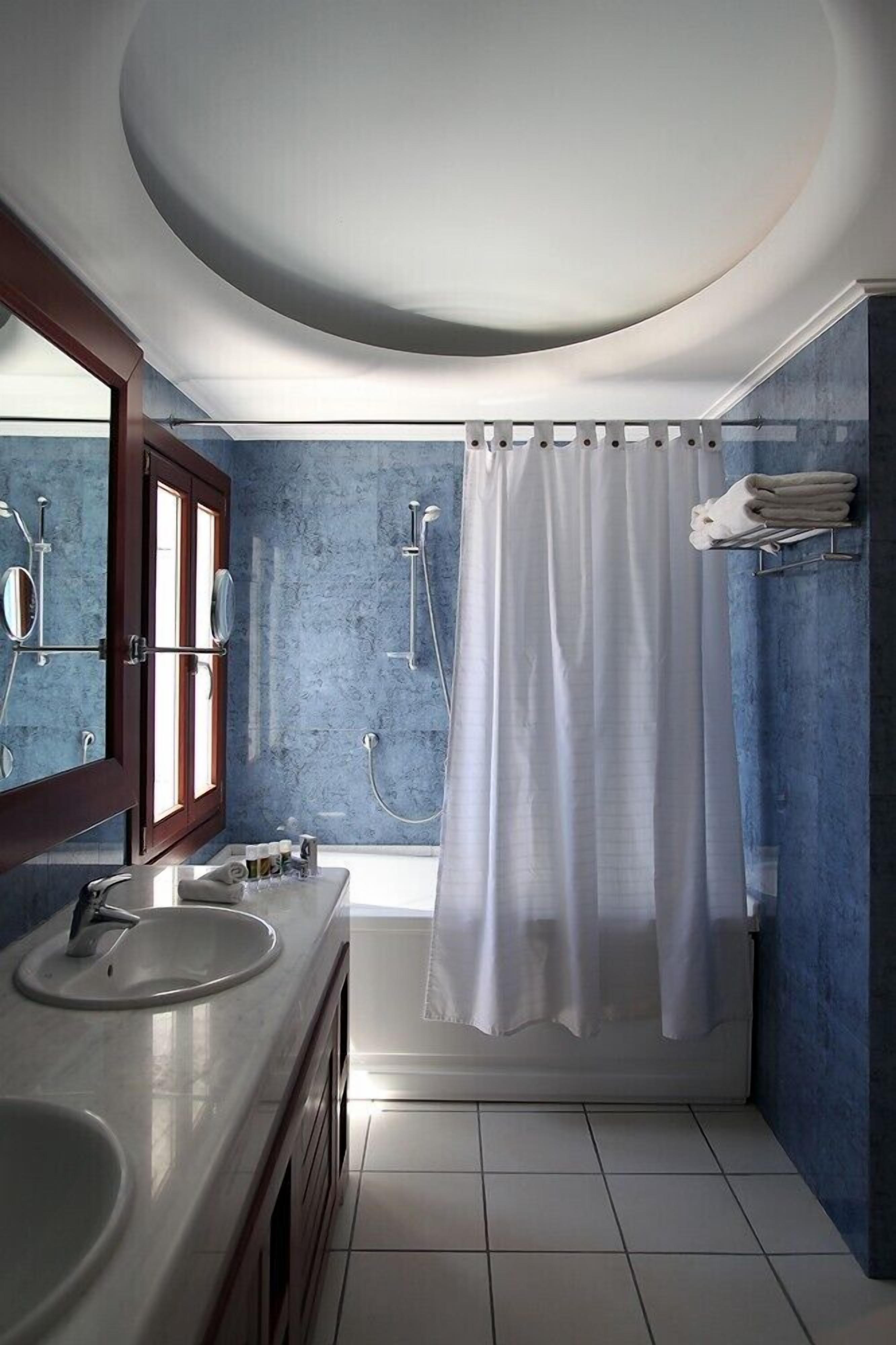 A bathroom with a double sink, a shower, a large mirror, and a window with curtains.