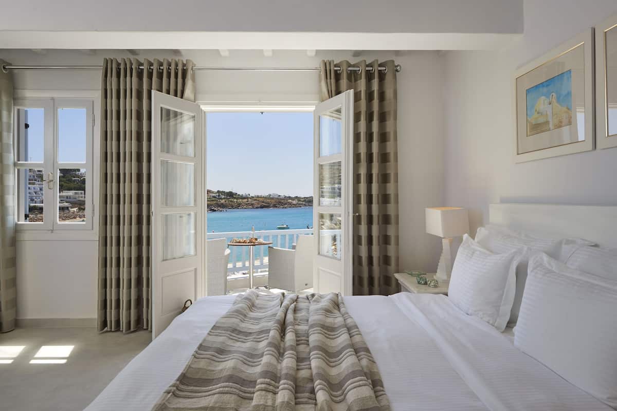 A hotel room with a bed, a nightstand with a lamp, a view of the sea, and a balcony with a table and chairs.