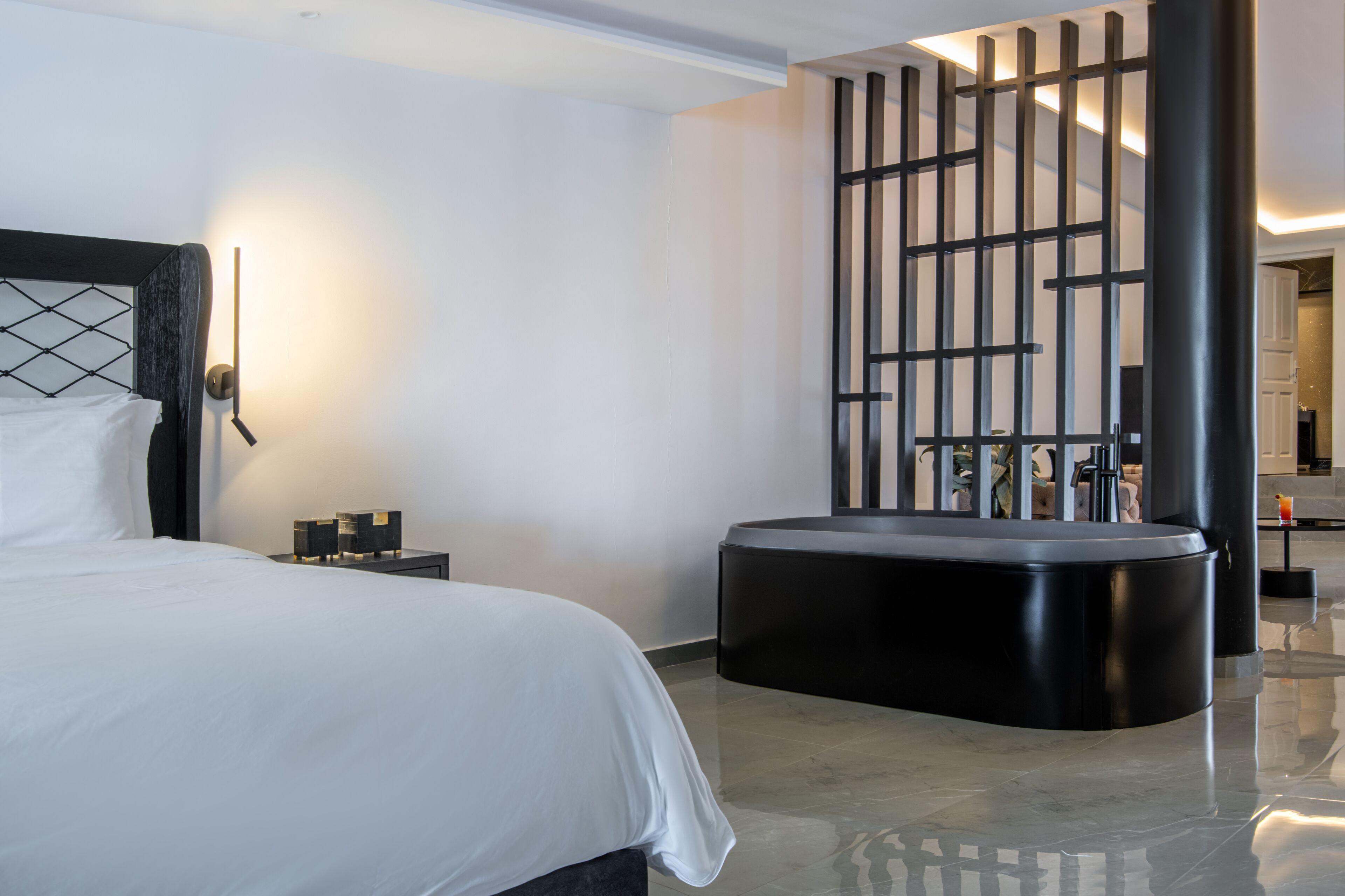 A modern hotel room with a large bed, a black ottoman, a nightstand with a lamp, and a decorative black partition.