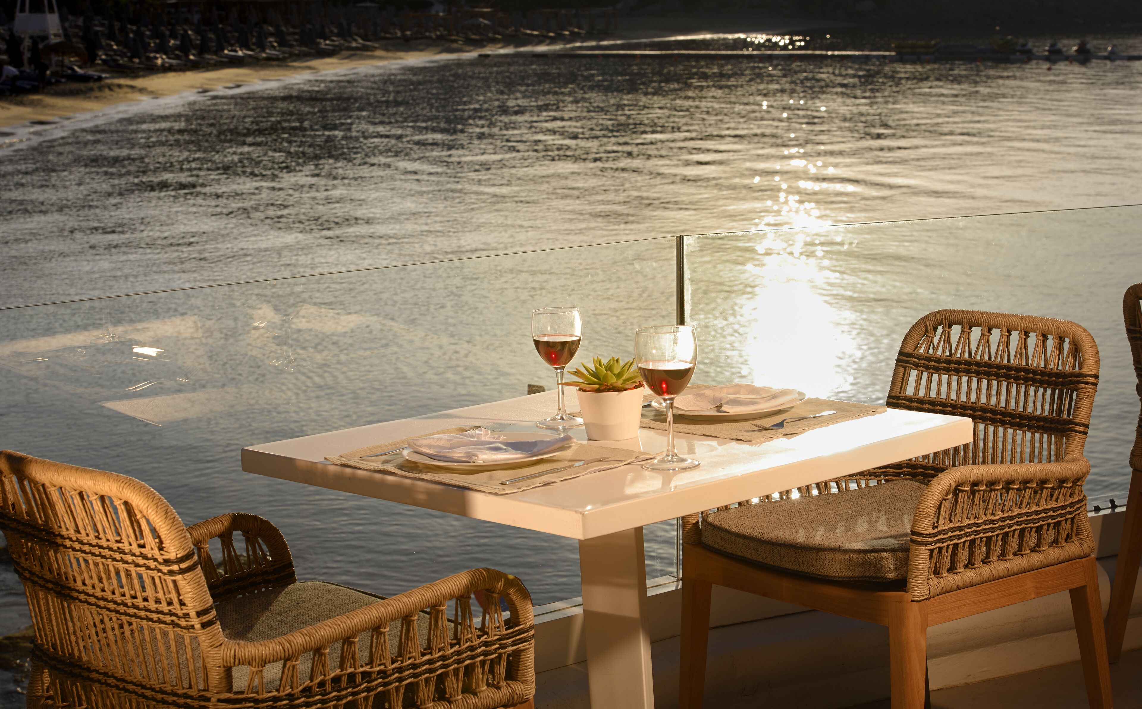 A table set for two with wine glasses and a small plant, overlooking a calm body of water.