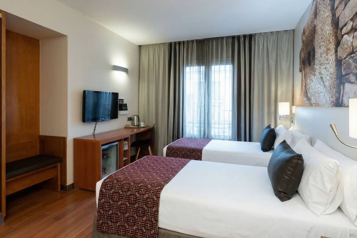 A hotel room with two beds, a desk, a television, and a large window with curtains.