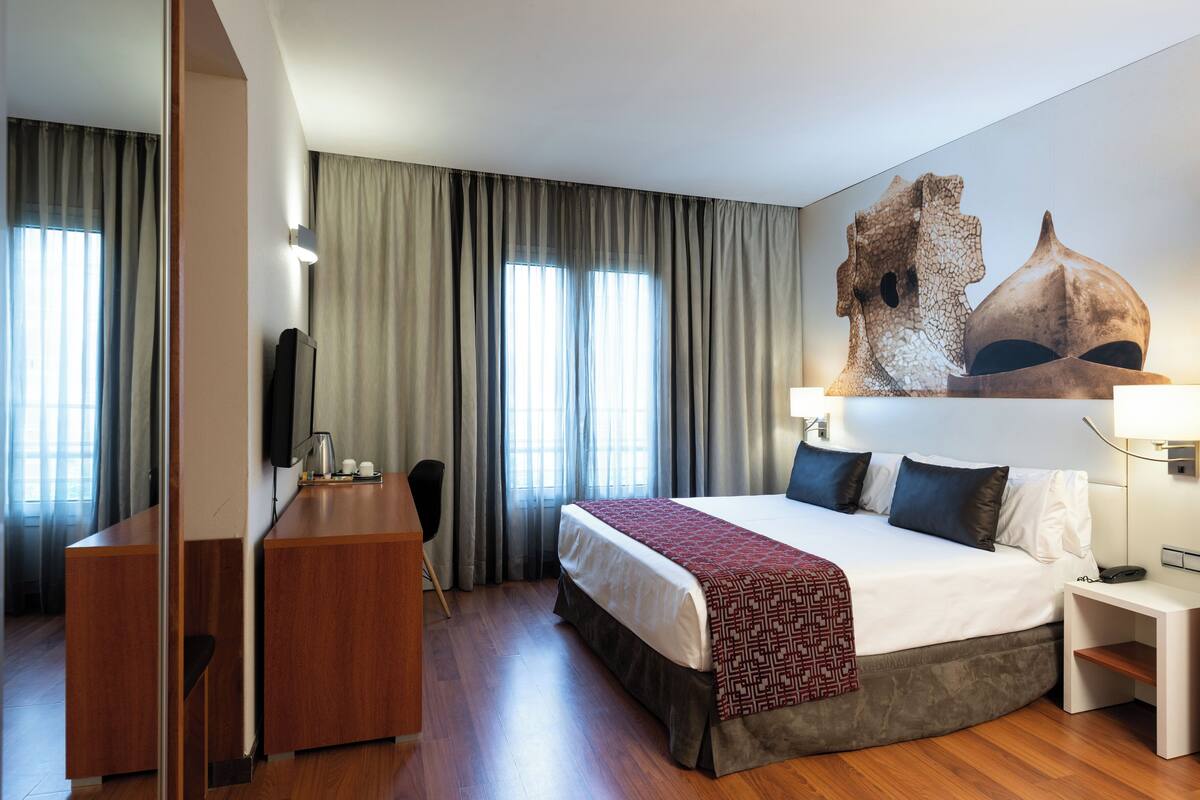 A hotel room with a bed, a desk, a chair, a large window with curtains, and a wall-mounted artwork.