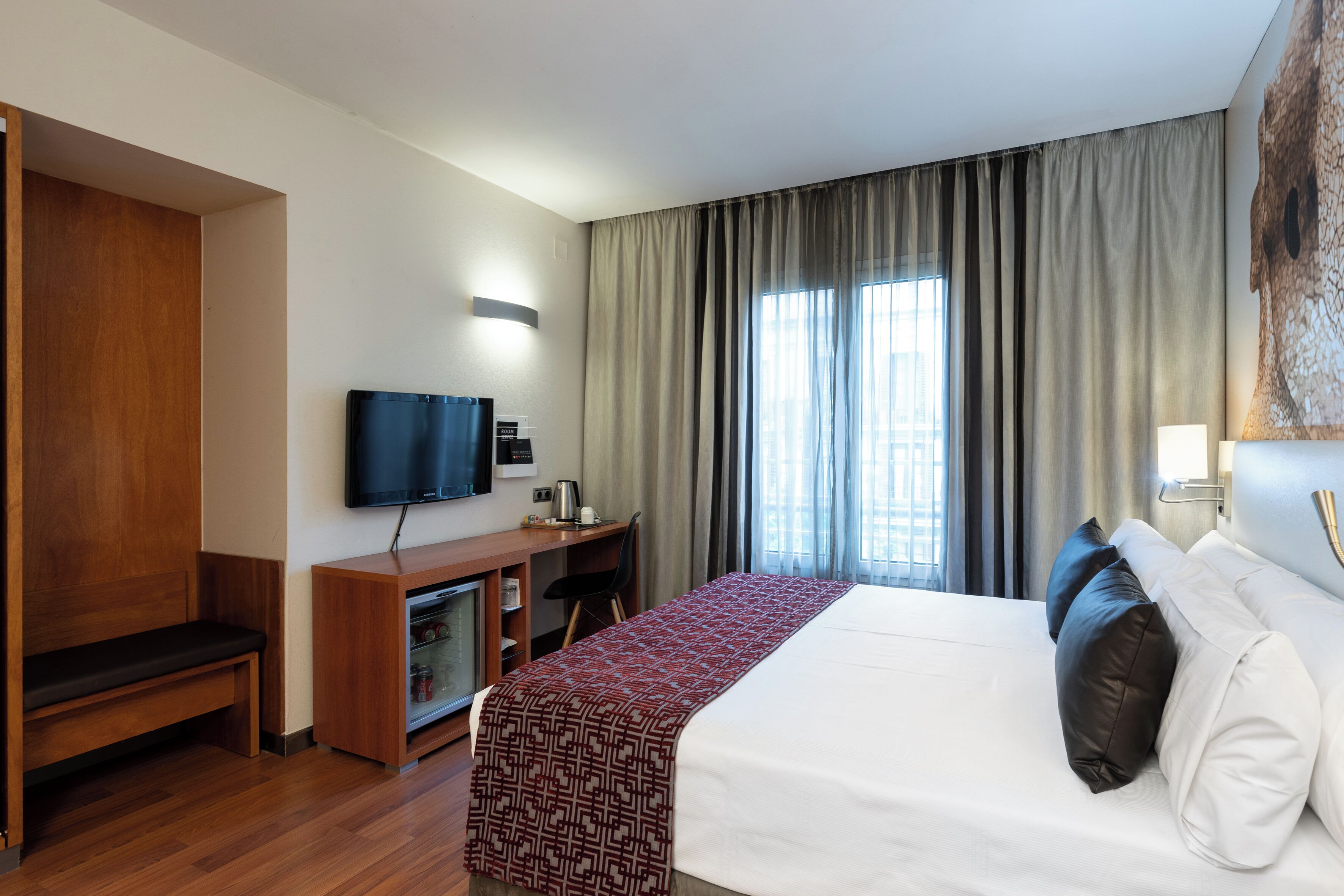 A hotel room with a bed, a desk with a television, a bench, and a window with curtains.