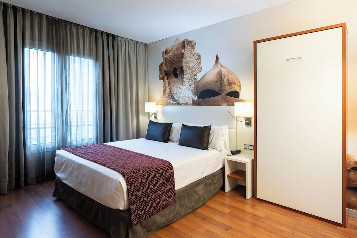 A hotel room with a large bed, a nightstand, a lamp, a wardrobe, and a window with curtains.