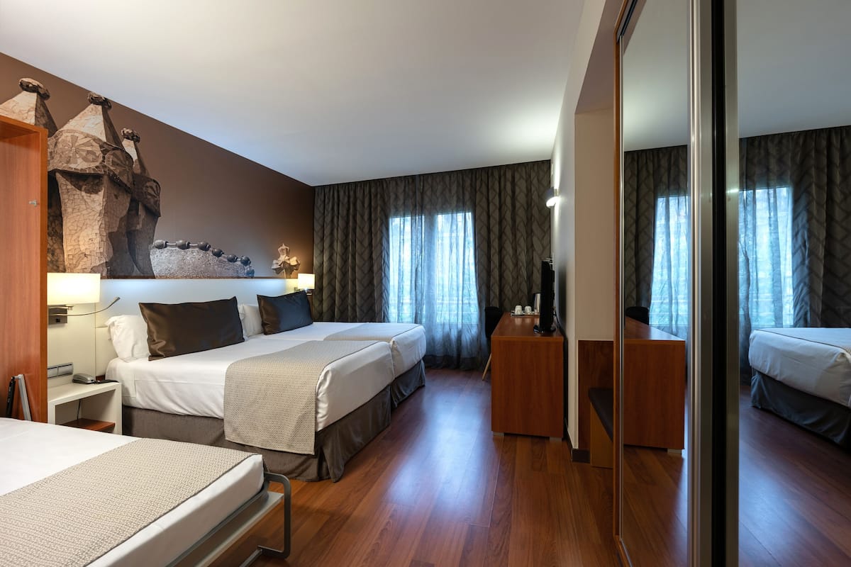 A hotel room with two beds, a desk, a mirror, and a large window with curtains.