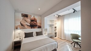 Una habitación de hotel moderna con una cama grande, un área de descanso con una silla, un armario y un amplio ventanal con cortinas.