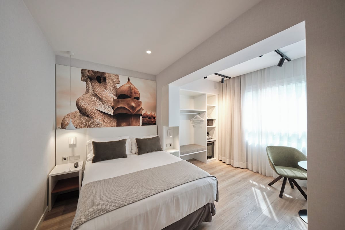 A modern hotel room with a large bed, a seating area with a chair, a wardrobe, and a large window with curtains.
