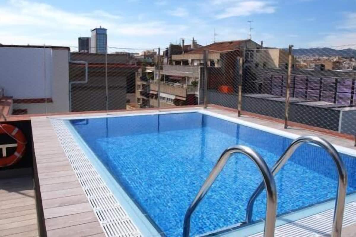 A rooftop swimming pool with metal railings, a lifebuoy, and a view of buildings and mountains.