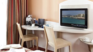 A hotel room with a desk, two chairs, a coffee maker, a television displaying a coastal view, and a small table with a cup. - Best Western Astoria (Antibes)