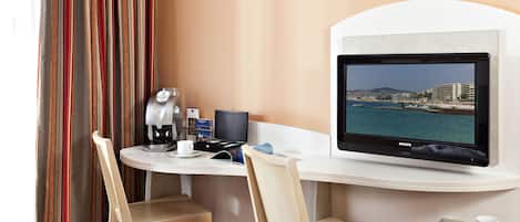 A hotel room with a desk, two chairs, a coffee maker, a television displaying a coastal view, and a small table with a cup.