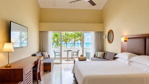 A hotel room with a bed, a desk, a chair, a table, and a balcony with a view of palm trees and the ocean.