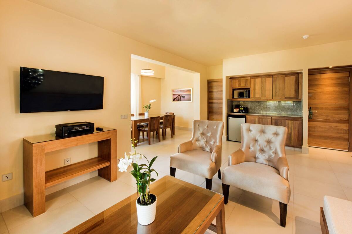 A modern living room with a flat-screen TV, a wooden coffee table, two beige armchairs, and a dining area with a wooden table and chairs.