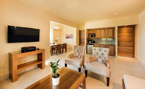 A modern living room with a flat-screen TV, a wooden coffee table, two beige armchairs, and a dining area with a wooden table and chairs.