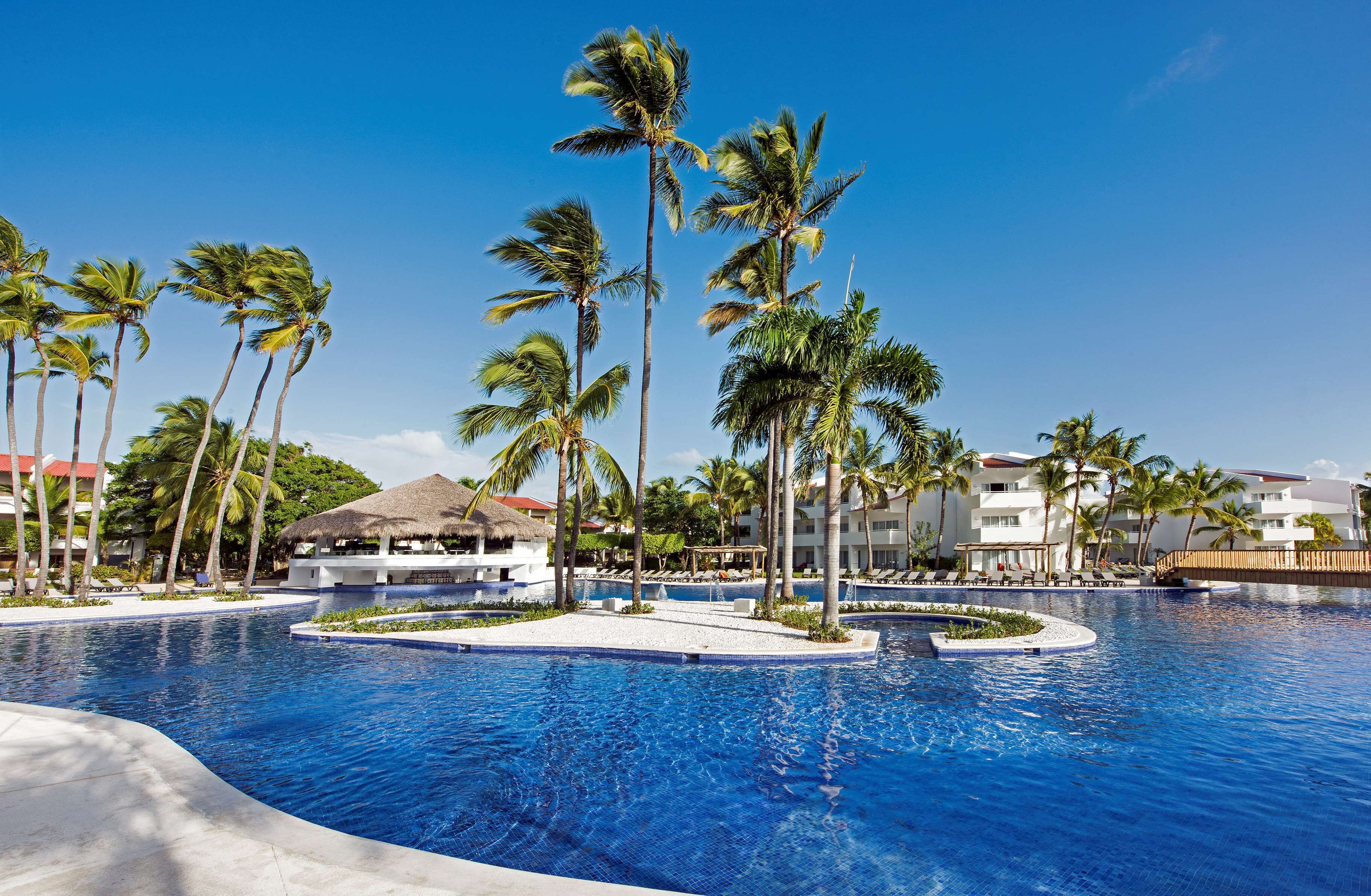 Piscina de resort con palmeras, una cabaña con techo de paja y un edificio de varios pisos al fondo.