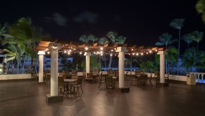 An outdoor patio area with tables and chairs, palm trees, and a pool in the background.