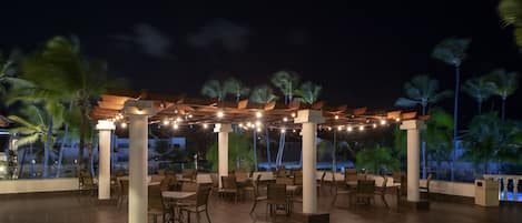An outdoor patio area with tables and chairs, palm trees, and a pool in the background.