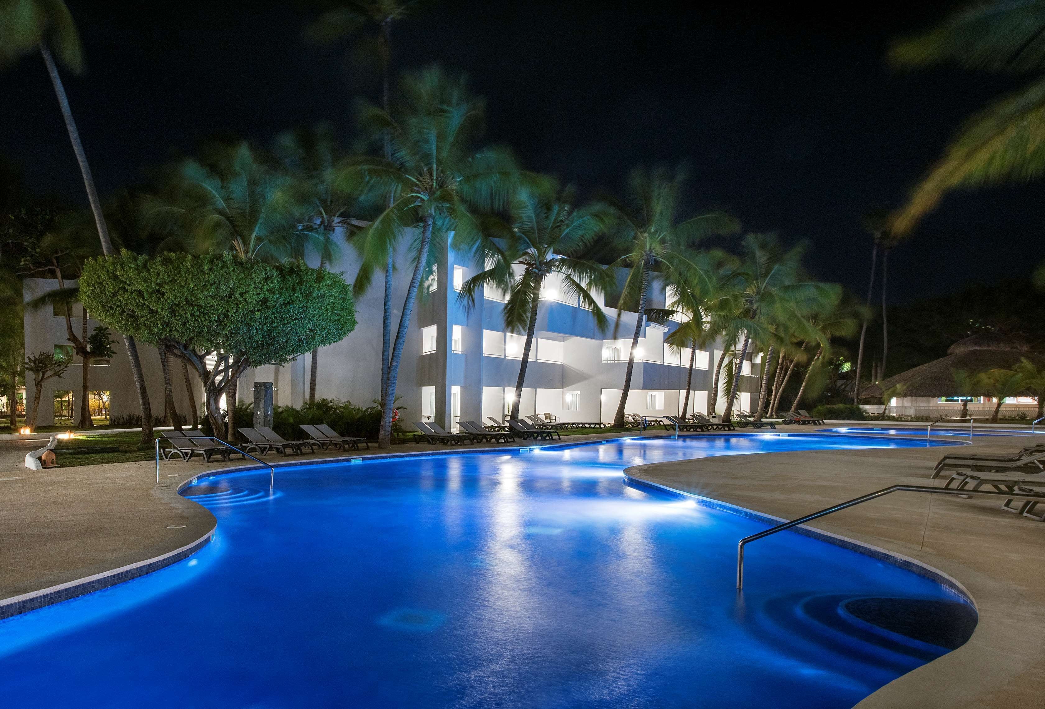 Una piscina con sillones de descanso y palmeras, rodeada por un edificio con ventanas iluminadas.