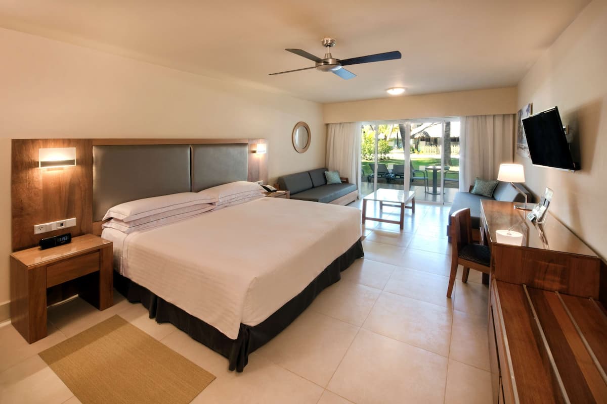 A hotel room with a large bed, a desk, a chair, a TV, and a balcony with outdoor seating.