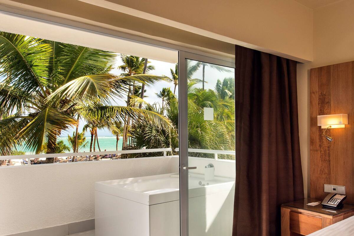 A hotel room with a glass shower enclosure, a wooden headboard, a bedside telephone, and a view of palm trees and the ocean.